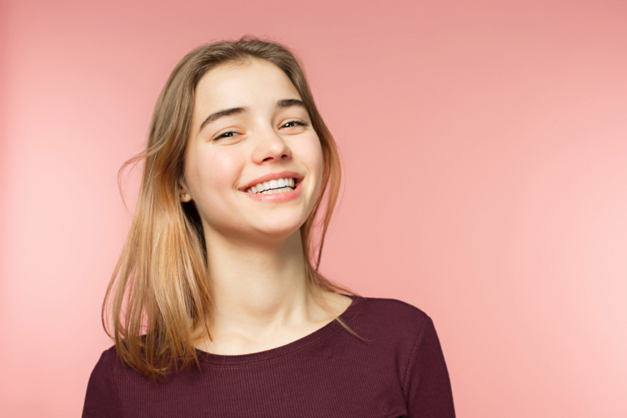 Teen girl with long hair and a bright, confident smile against a pink background, showing the straight teeth Invisalign Birmingham can create at Orthodontic Associates with Dr. Gregory Hummon and Dr. Thomas Gebeck Jr.
