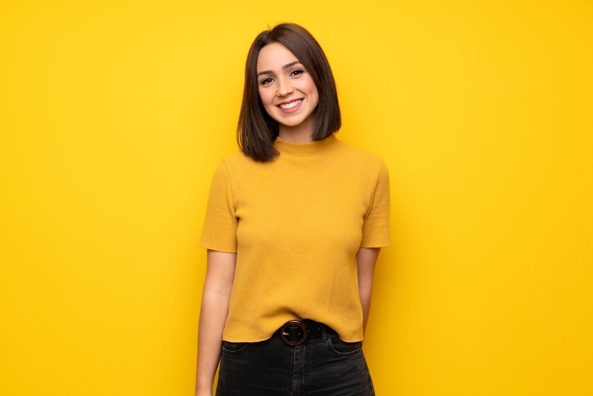 Smiling young woman with straight teeth in a yellow sweater against a yellow background, showing results of Invisalign Birmingham treatment with Dr. Gregory Hummon and Dr. Thomas Gebeck Jr. at Orthodontic Associates